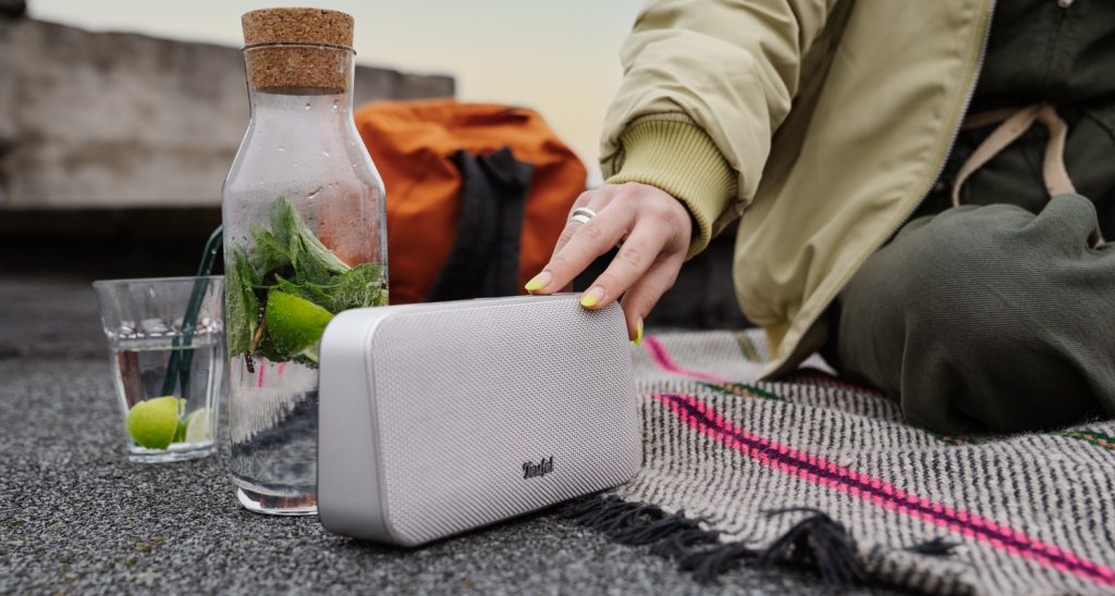 Głośnik MOTIV GO ustawiony na kocu na tarasie obok wody.